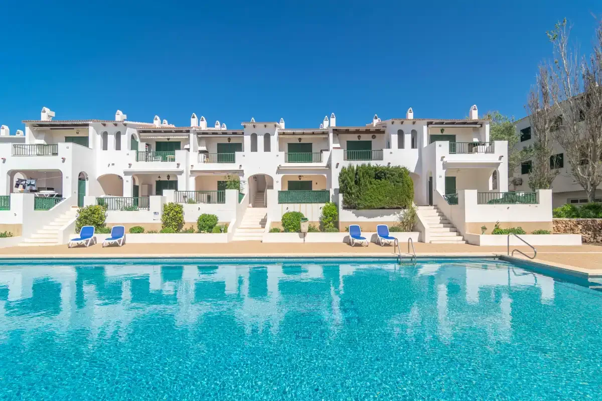 Una piscina frente a edificios blancos de apartamentos bajo un cielo azul despejado.
