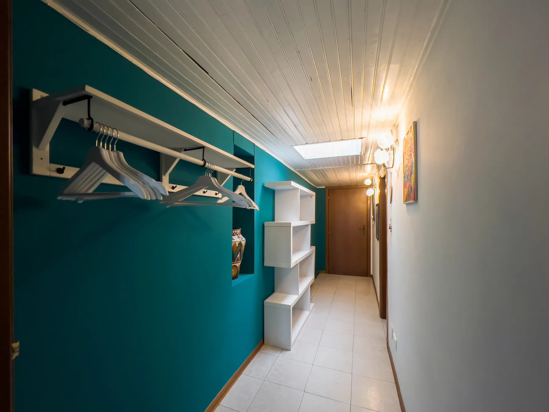 Un pasillo estrecho con estantes abiertos y una barra para ropa junto a una pared verde azulado.