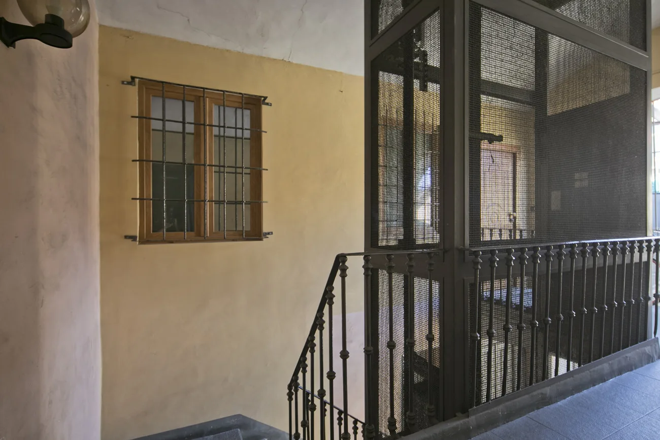 Un pasillo interior con suelo de baldosas, una ventana enrejada y una barandilla metálica.