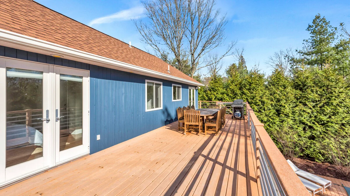 A wooden deck runs alongside a blue house, with outdoor seating and a railing.