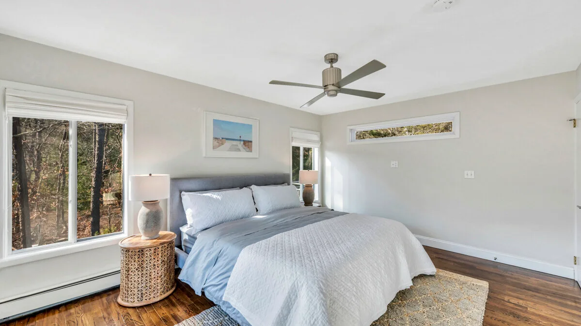 A bright bedroom with a neatly made bed and windows overlooking trees.