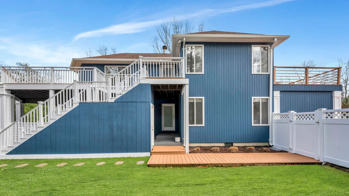 A blue house with white exterior stairs and a wooden deck beside a lawn.