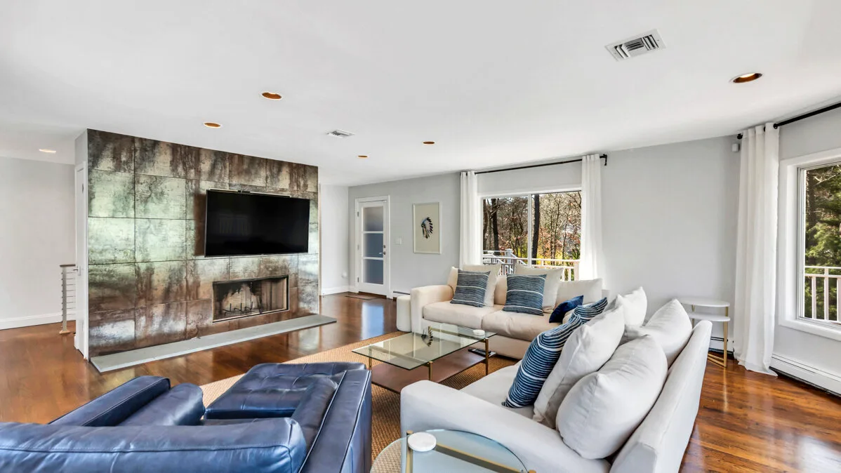 A bright living room with a television above a fireplace and seating by the windows.
