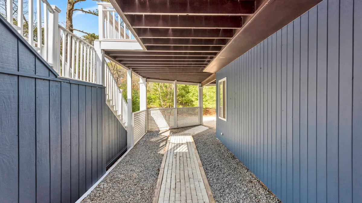A narrow outdoor passage with a gravel surface and a wooden walkway beneath a raised deck.