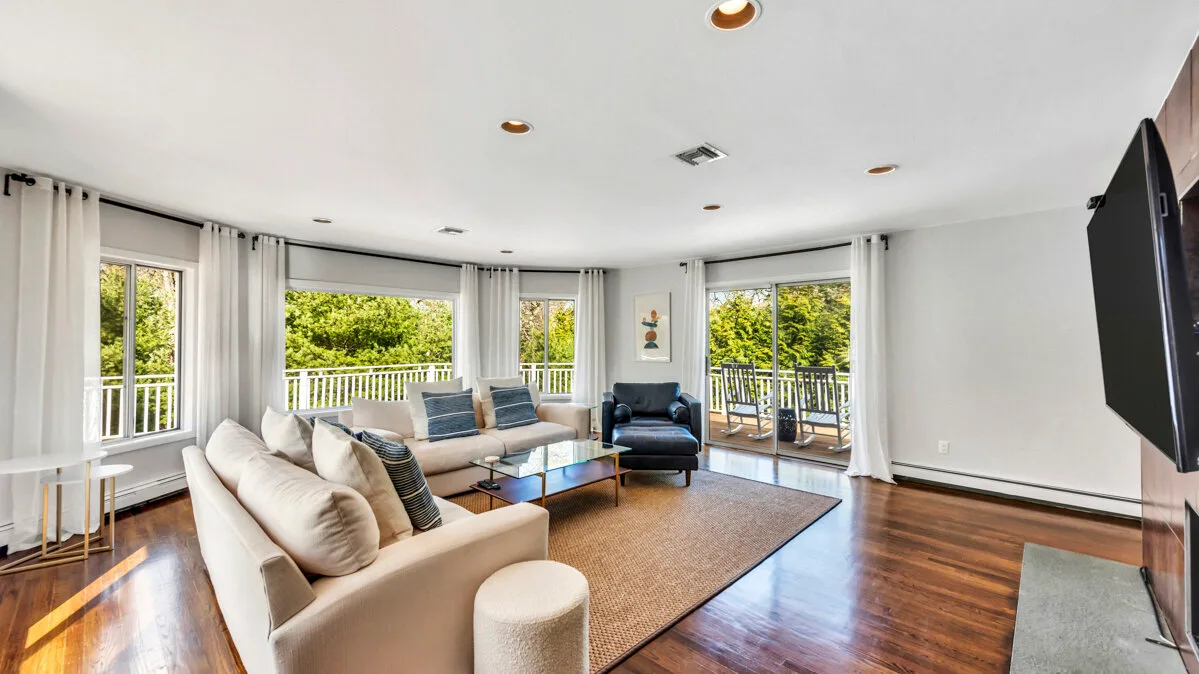 A bright living room with seating, a rug, and wide windows overlooking greenery.