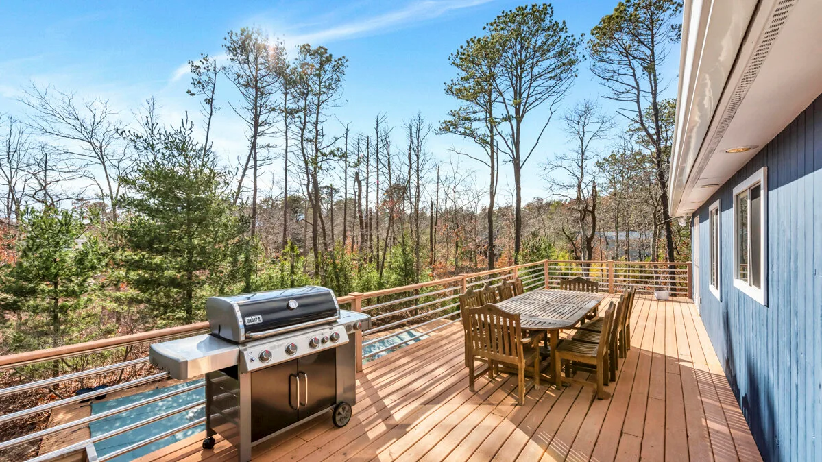 A wooden deck has an outdoor grill and a dining table beside the house.