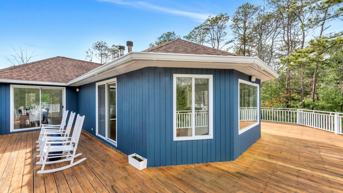 A blue house opens onto a broad wooden deck with white railings and outdoor seating.
