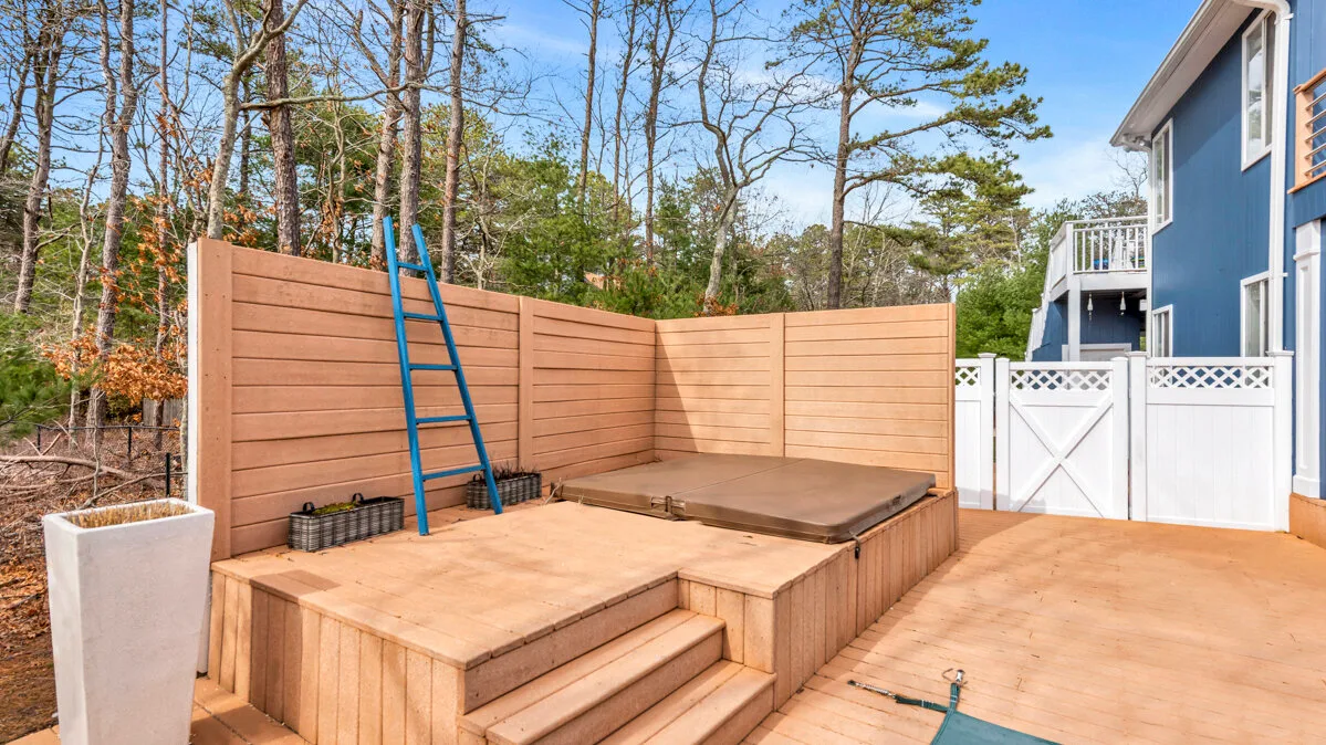 A wooden deck with a raised hot tub and privacy fencing beside a blue house.