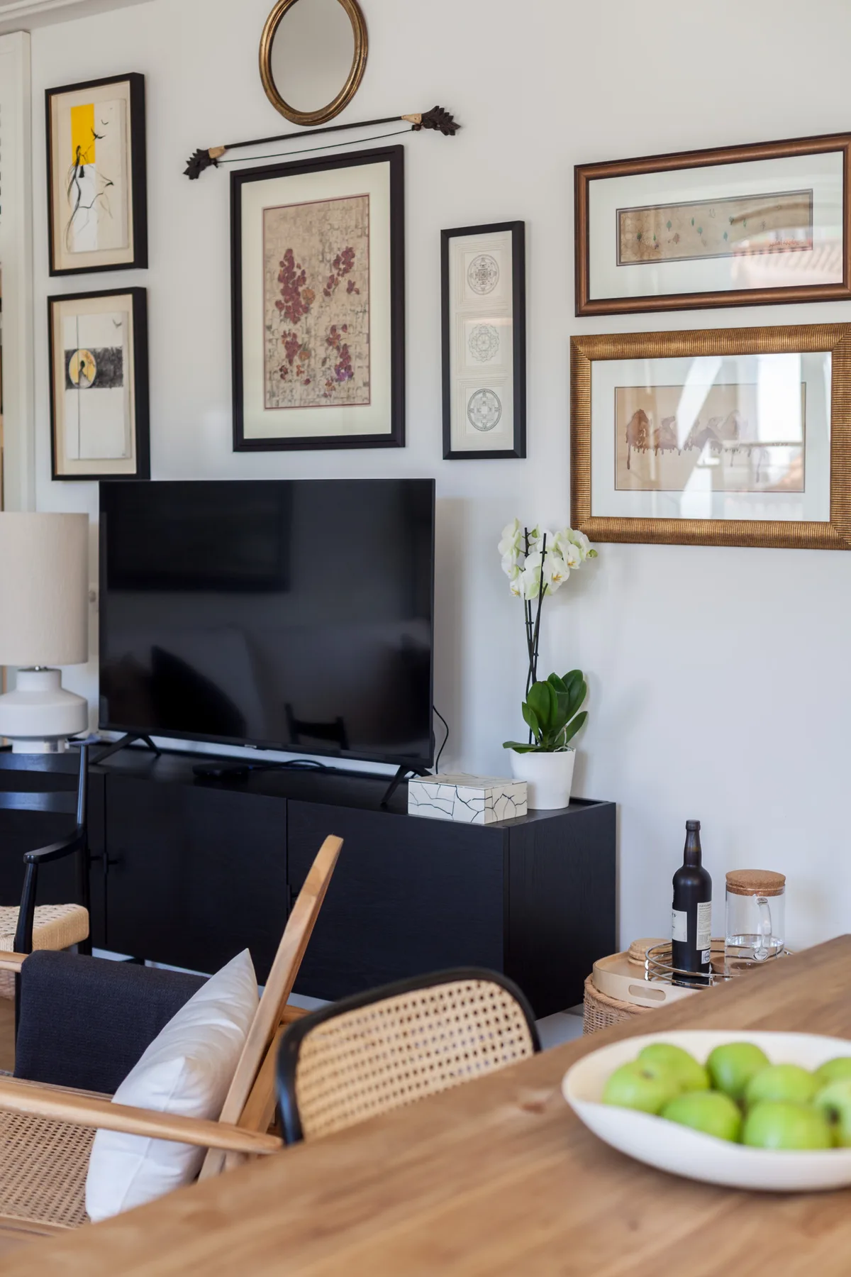 A dining area with a wooden table and seating faces a television beneath framed artwork.