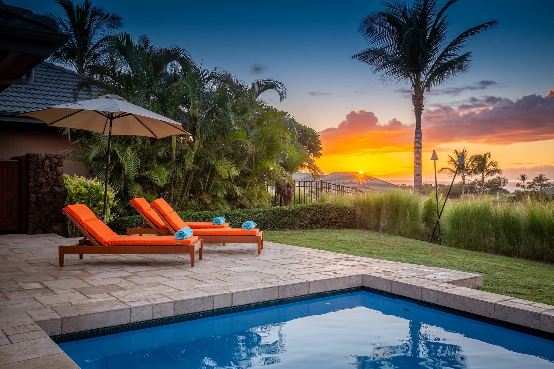 Un patio junto a la piscina con tumbonas naranjas mira hacia palmeras y el atardecer.
