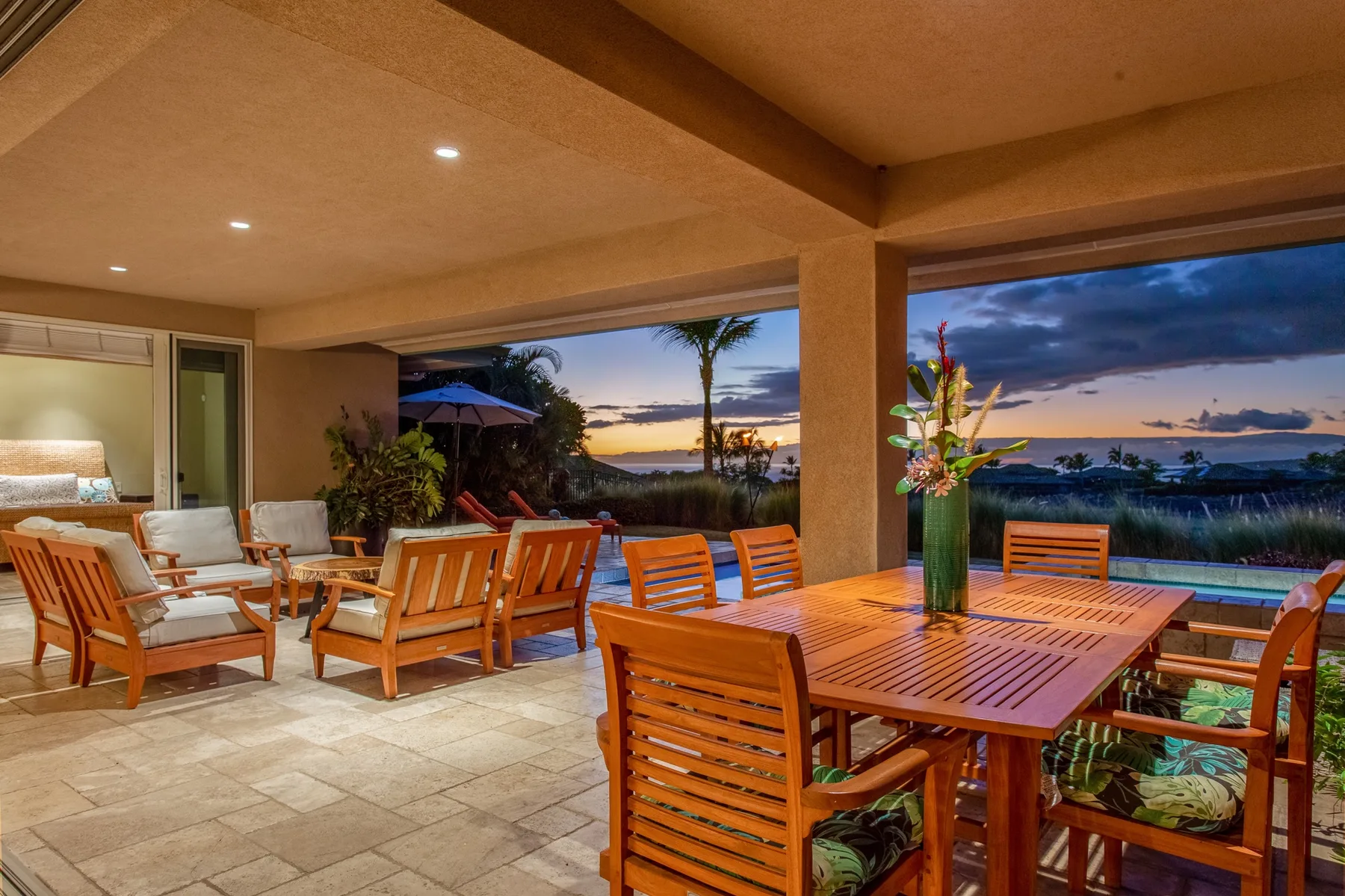 Un patio cubierto con asientos de madera y una mesa de comedor mira al océano al atardecer.