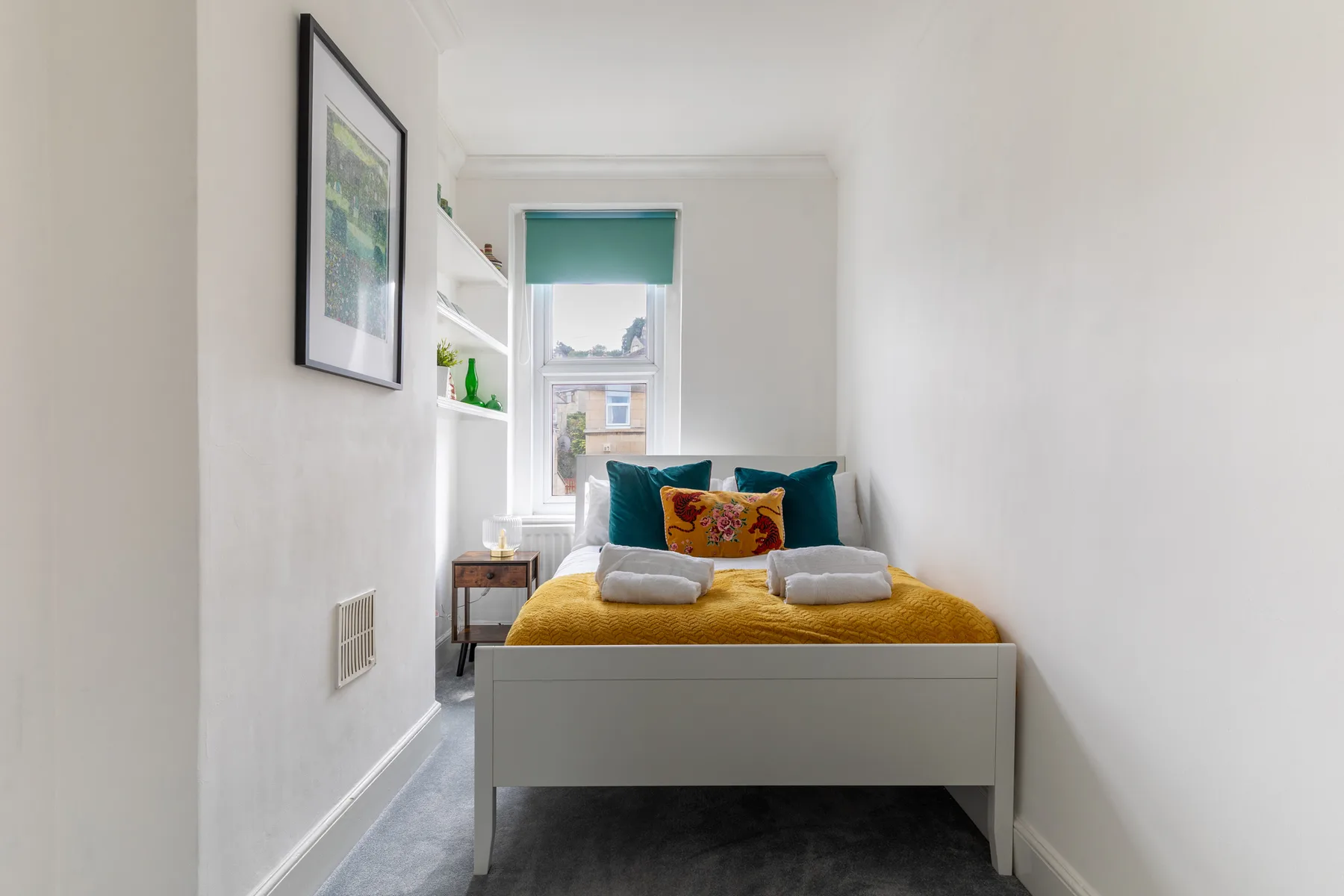 A narrow bedroom with a bed beneath a window and a teal roller blind.