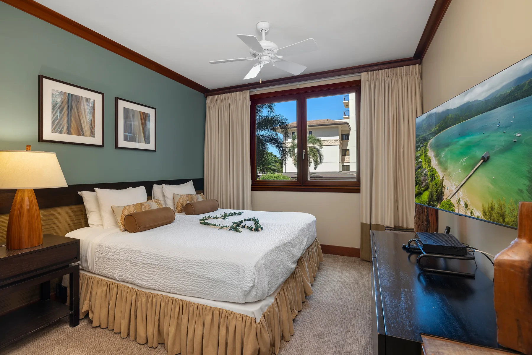 Un dormitorio con una cama bien tendida, cuadros enmarcados y una ventana con vista a palmeras.
