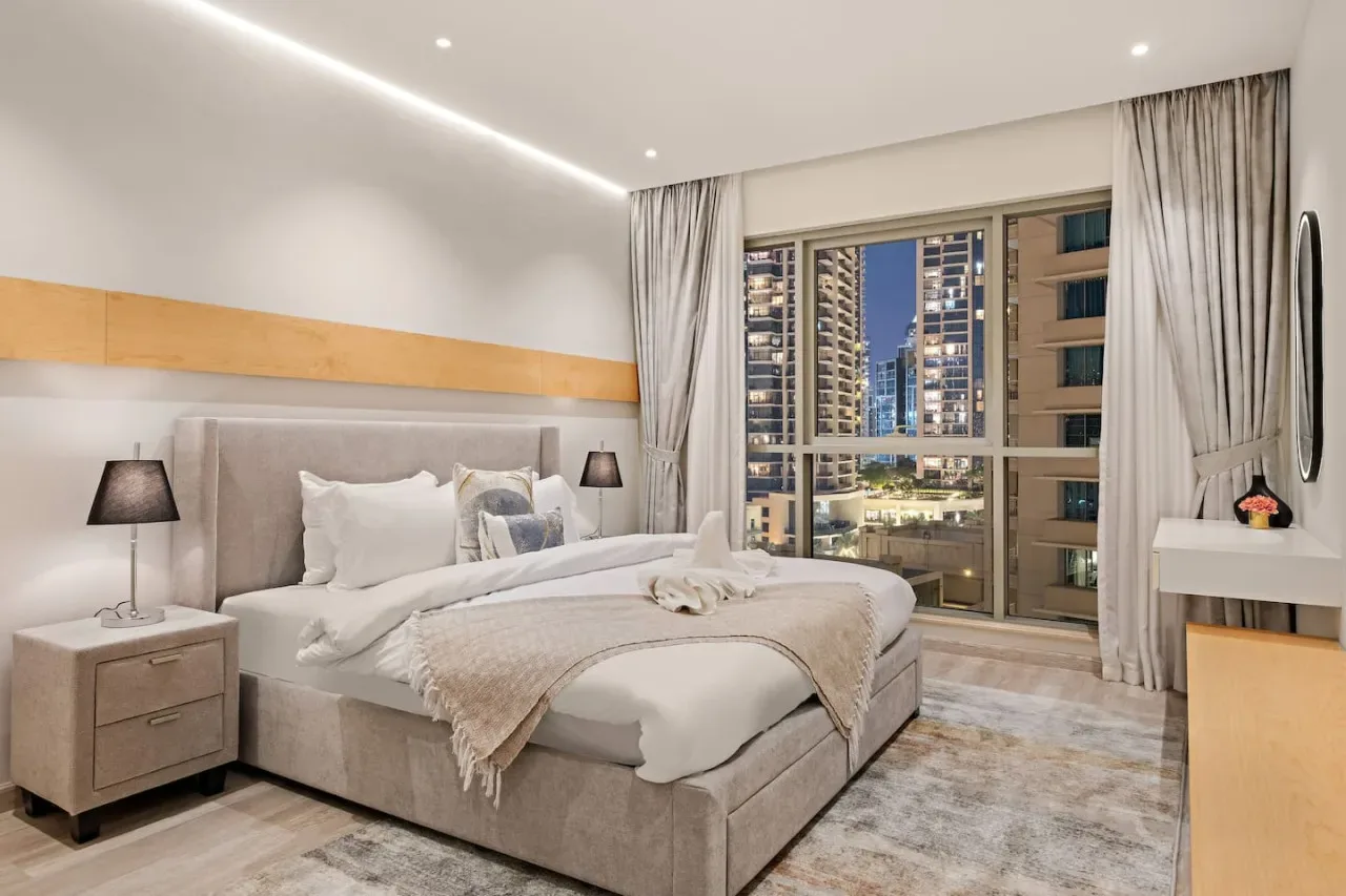 Un dormitorio con una cama grande, mesitas de noche y una ventana con vistas a edificios cercanos.