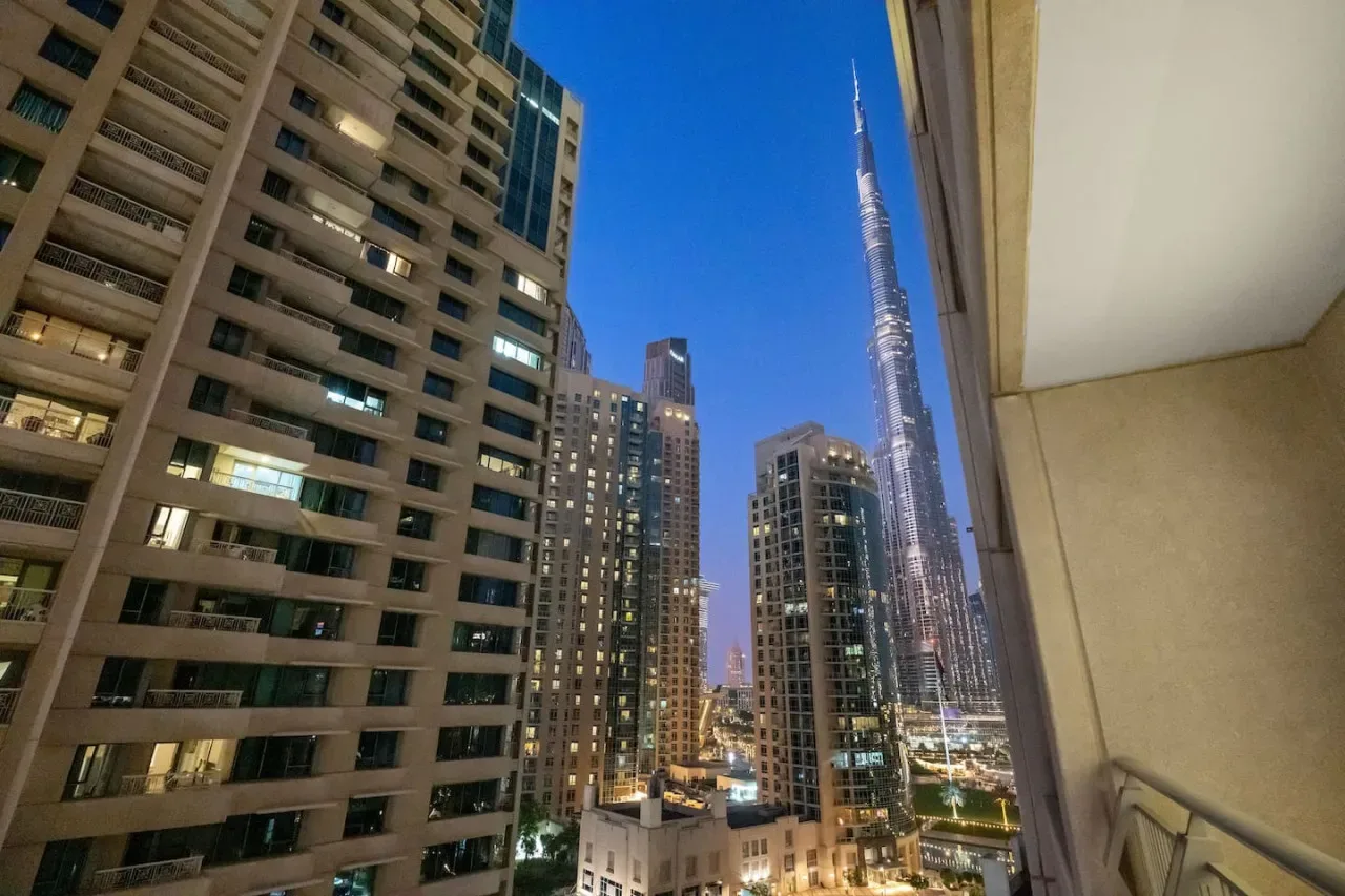 Vista nocturna de la ciudad desde un balcón, con edificios iluminados y una torre esbelta.