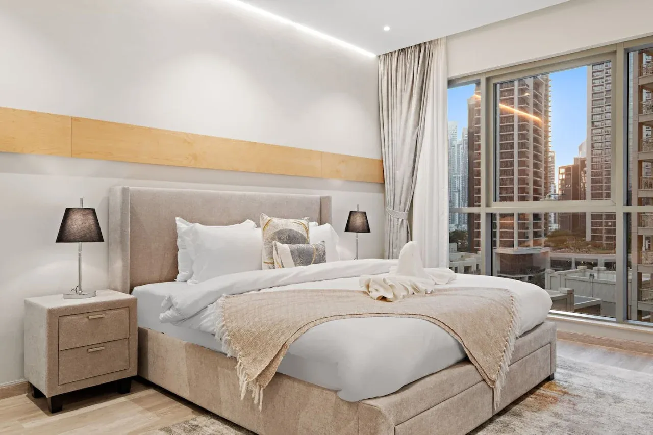 Un dormitorio con una cama grande junto a una ventana amplia con vistas a edificios cercanos.