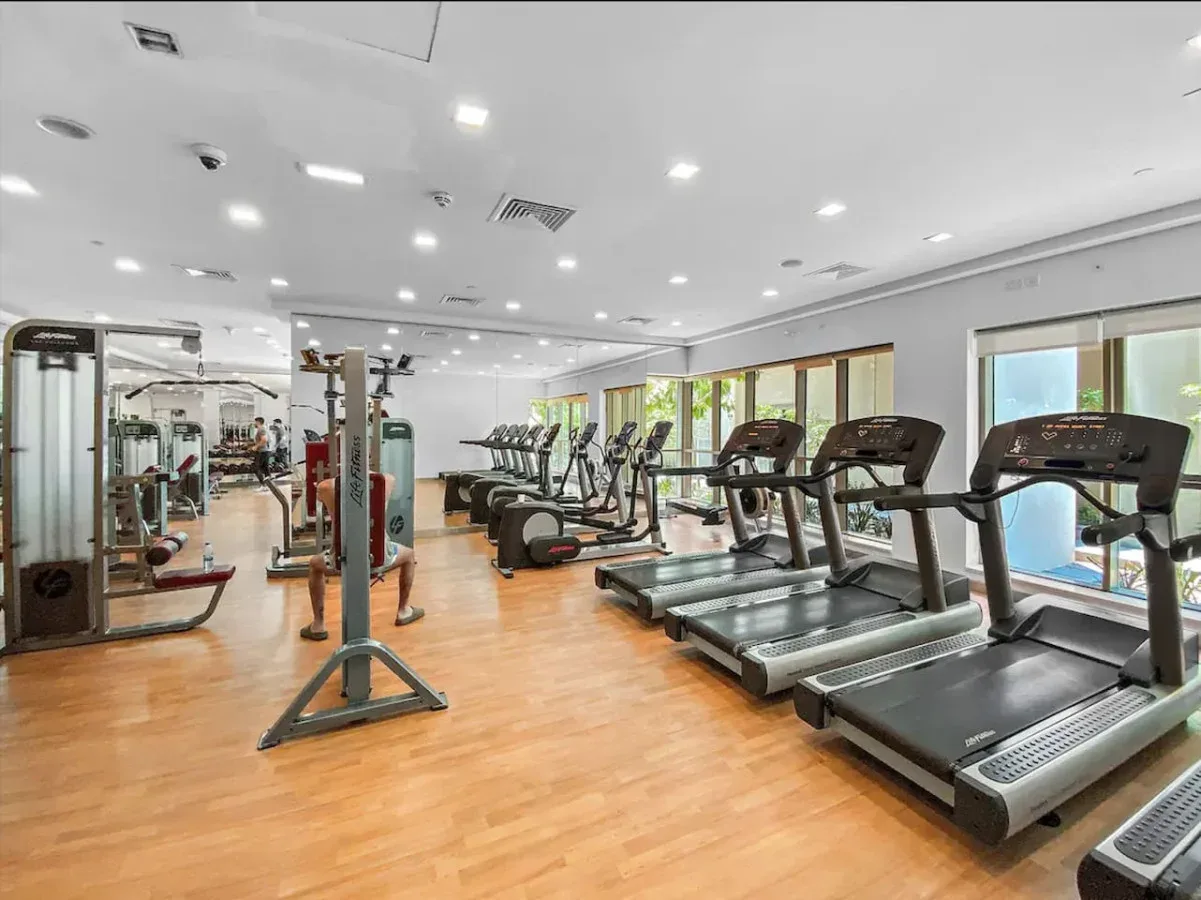 Sala de ejercicio con cintas de correr, máquinas y grandes ventanales.