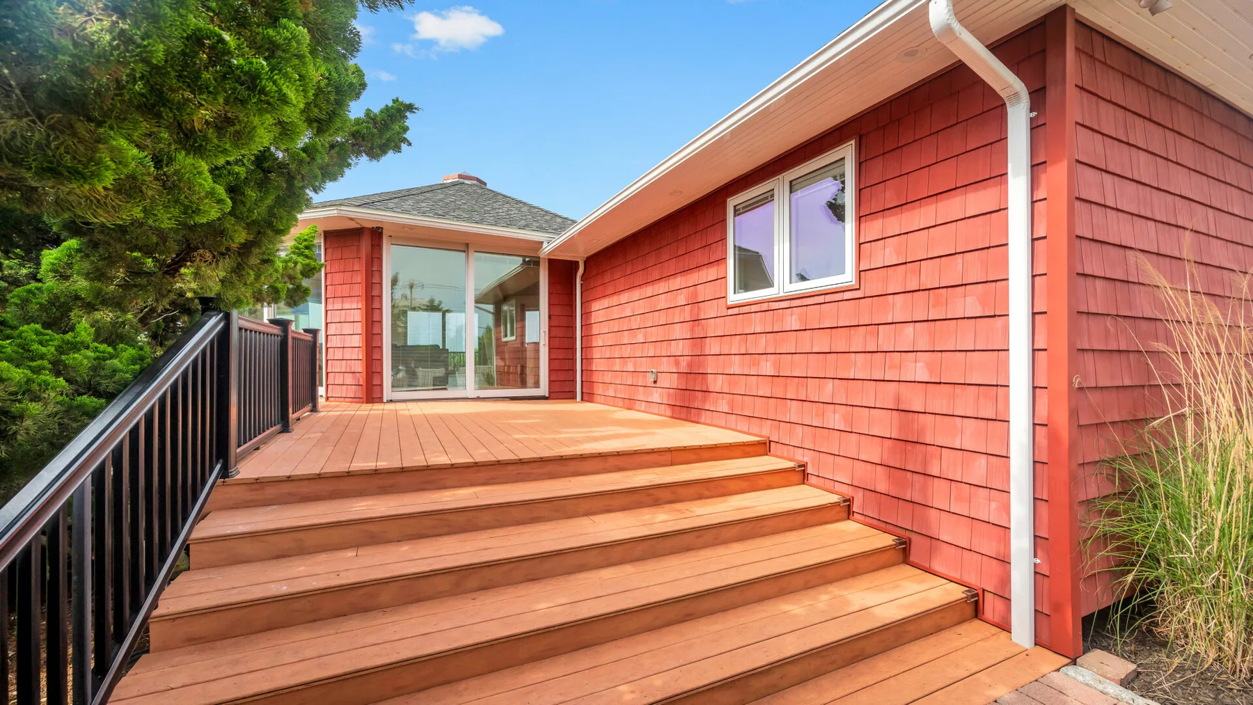 Una terraza de madera con escalones anchos conduce a una puerta de vidrio junto a una casa roja.