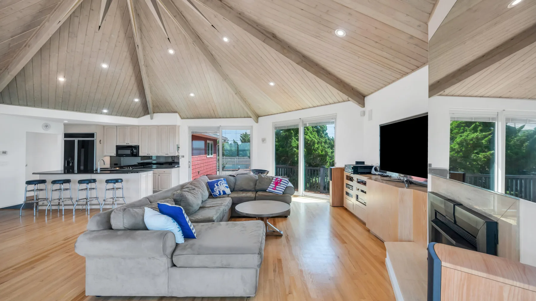 Una sala de estar de concepto abierto con zona de asientos gris, cocina y techo abovedado de madera.