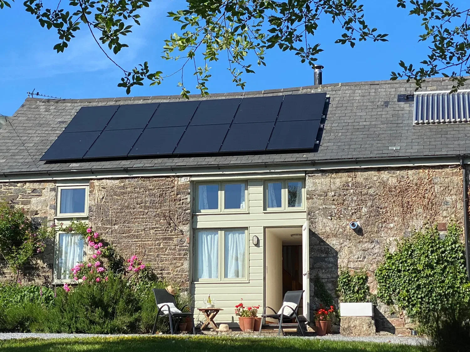 Una casa de campo de piedra con paneles solares en el tejado y dos sillas con una mesa pequeña en el exterior en The Stone Barn Cottage, en Holne