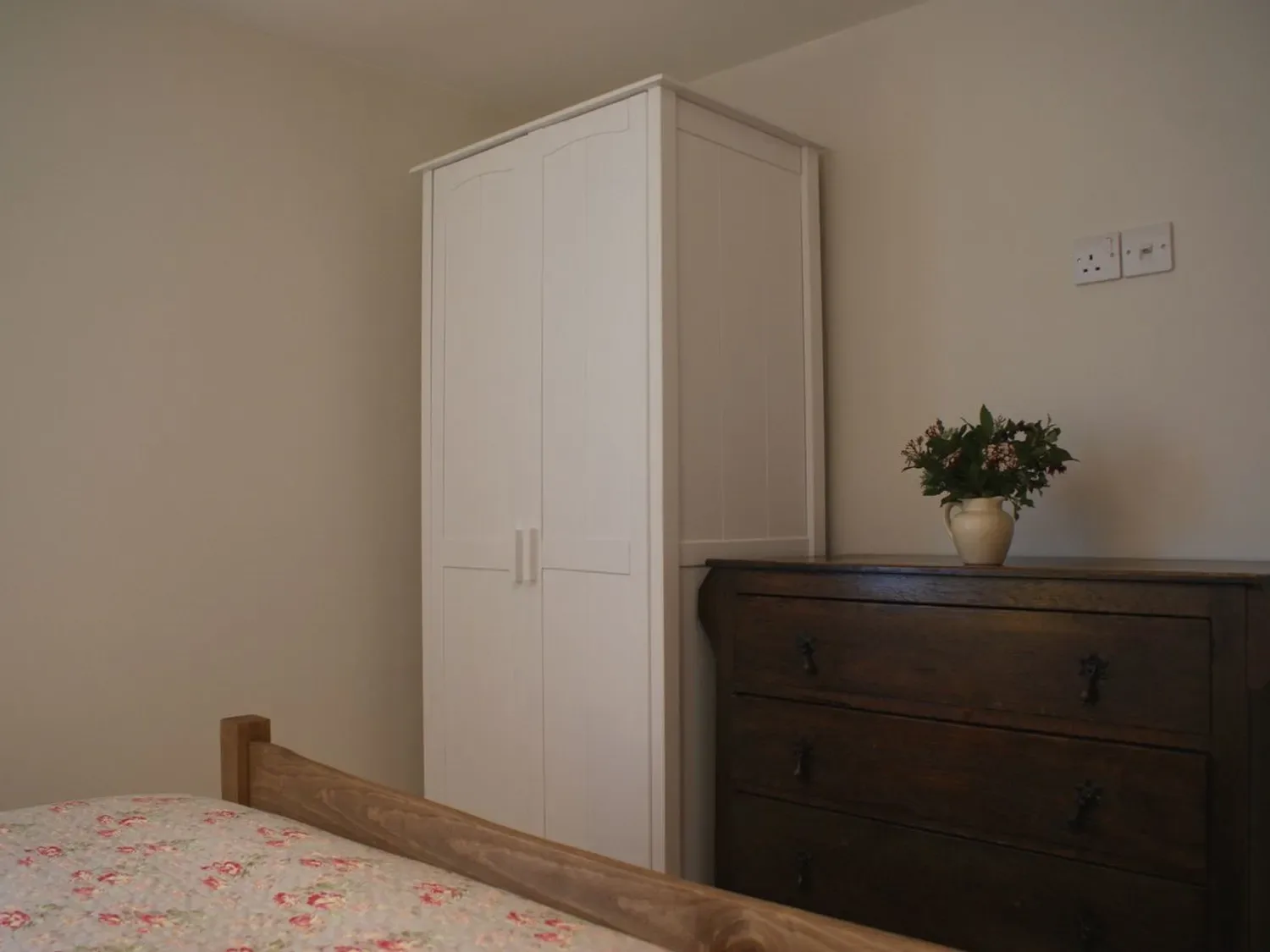Un dormitorio con un armario blanco, una cómoda oscura y ropa de cama floral.