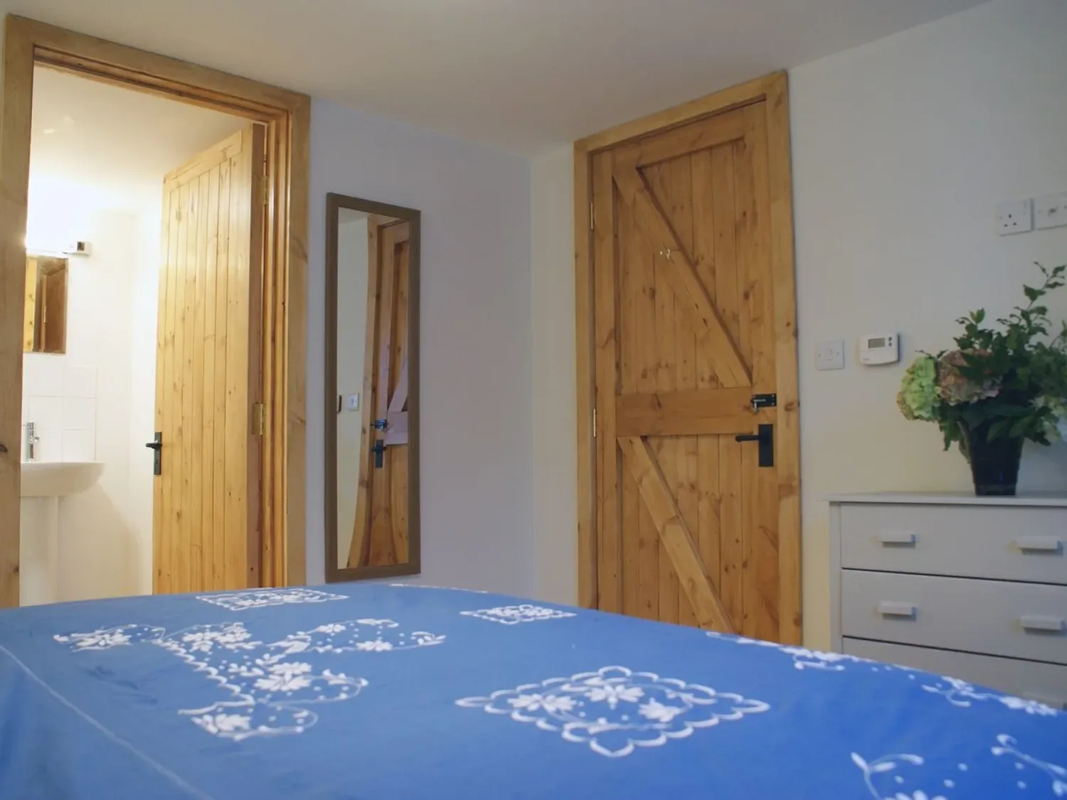 Un dormitorio con puertas de madera, una colcha azul estampada, una cómoda blanca y un espejo en la pared en The Stone Barn Cottage, en Holne