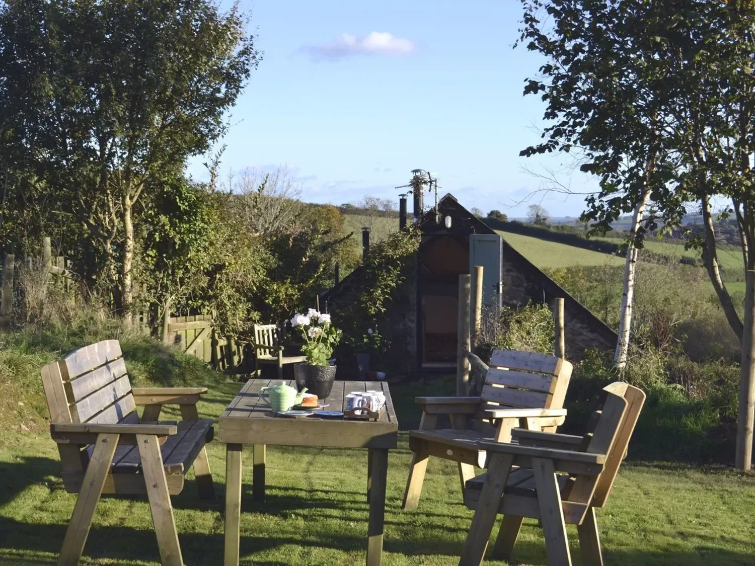Un jardín con sillas de madera y una mesa con una tetera y flores en el exterior de un edificio de piedra en The Stone Barn Cottage, en Holne