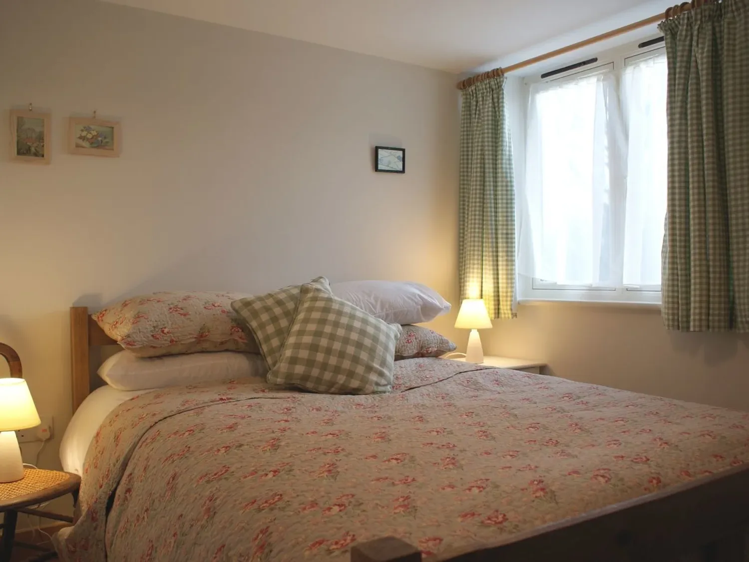 Un dormitorio con ropa de cama floral, cojines de cuadros, lámparas de mesilla y una ventana con cortinas.