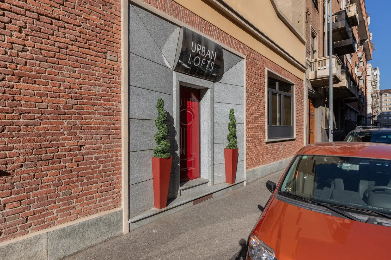 A red entrance door sits in a gray stone surround on a brick building beside a street.