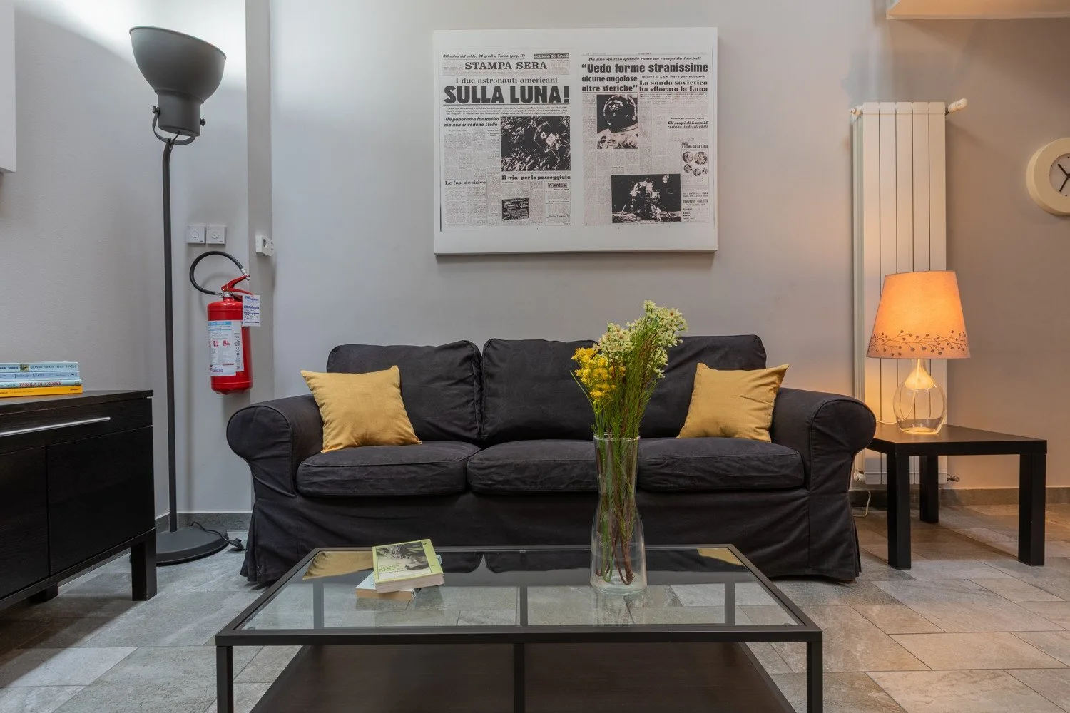 A living room with dark seating, a glass coffee table, and a framed newspaper display.