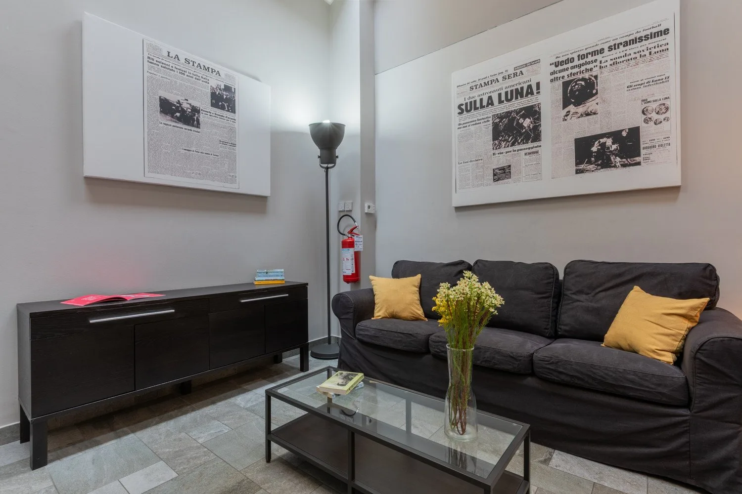 A living room with dark seating, framed newspaper pages, and a glass coffee table.