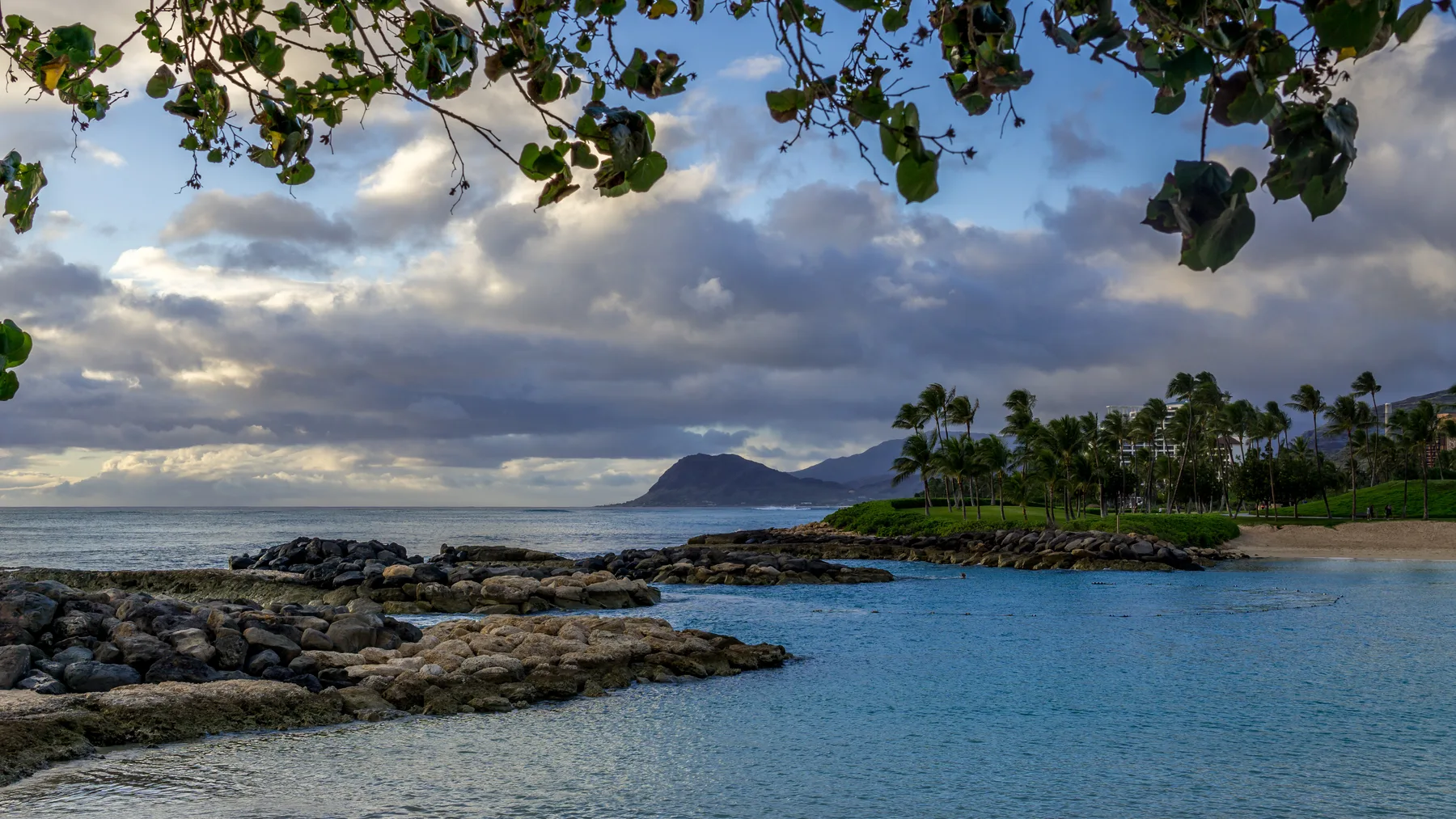 A rocky shoreline borders calm blue water beneath a cloudy sky.