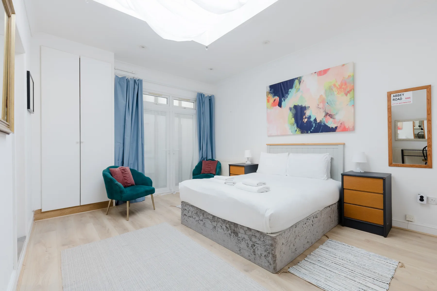 Un dormitorio luminoso con una cama blanca, cortinas azules y arte colorido en la pared.