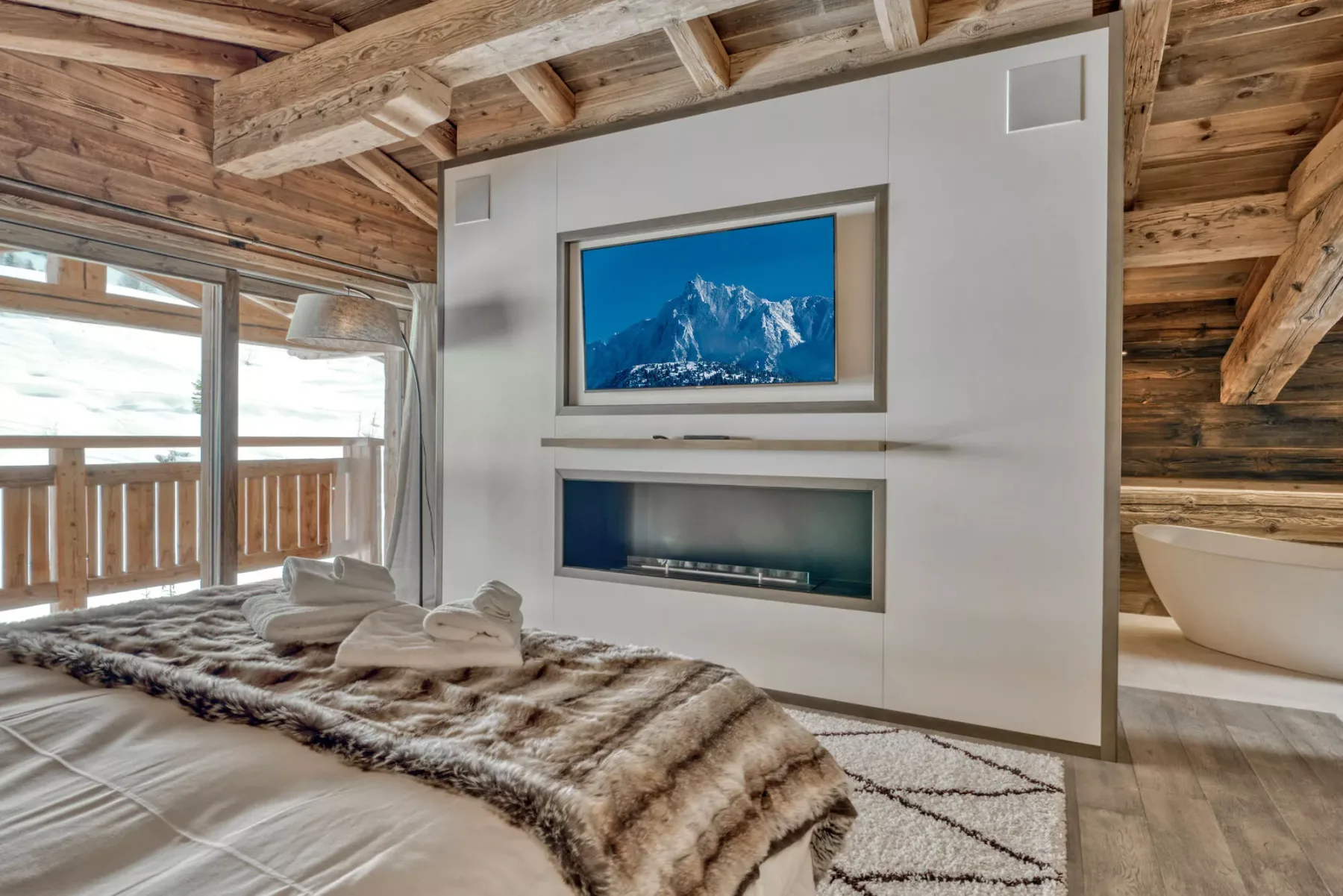 A bedroom features a bed with a fur throw, a wall-mounted television, and a freestanding bathtub.