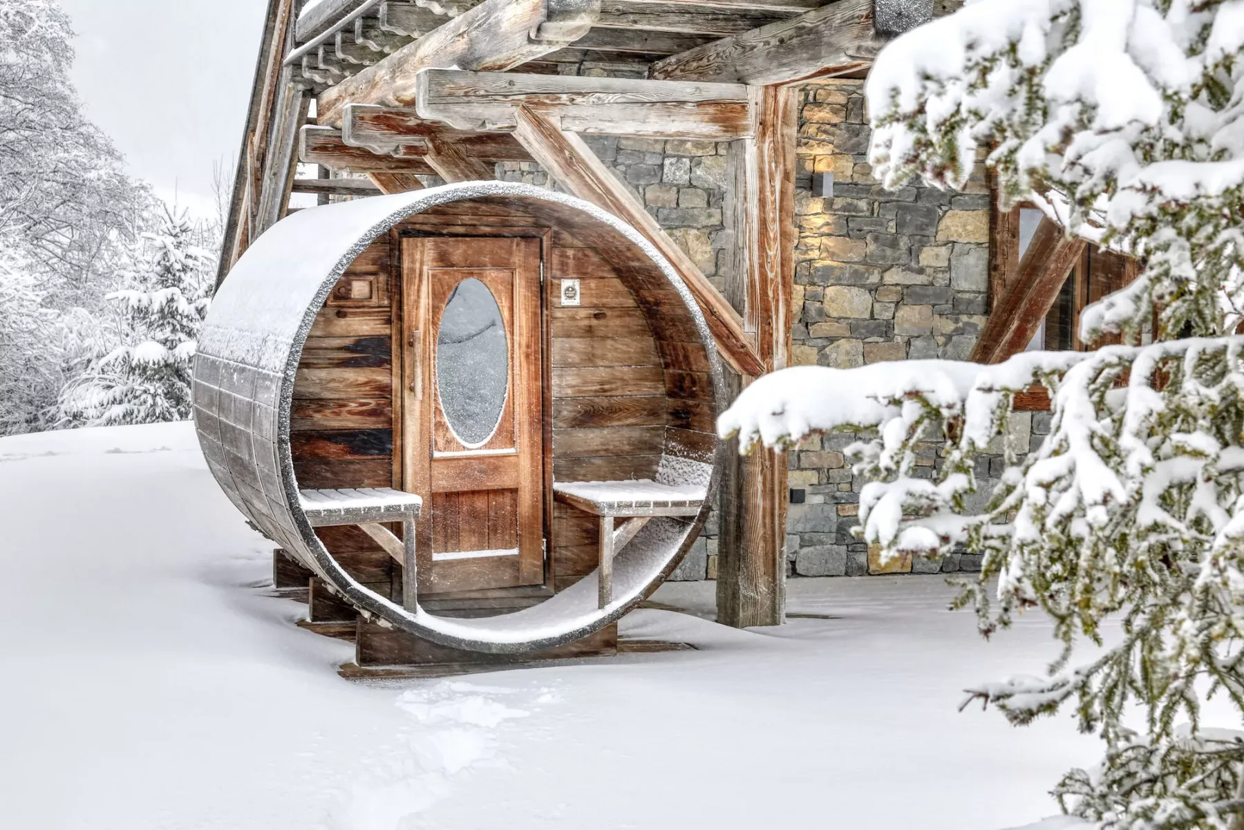 A wooden barrel-shaped sauna stands outside a stone building in the snow.