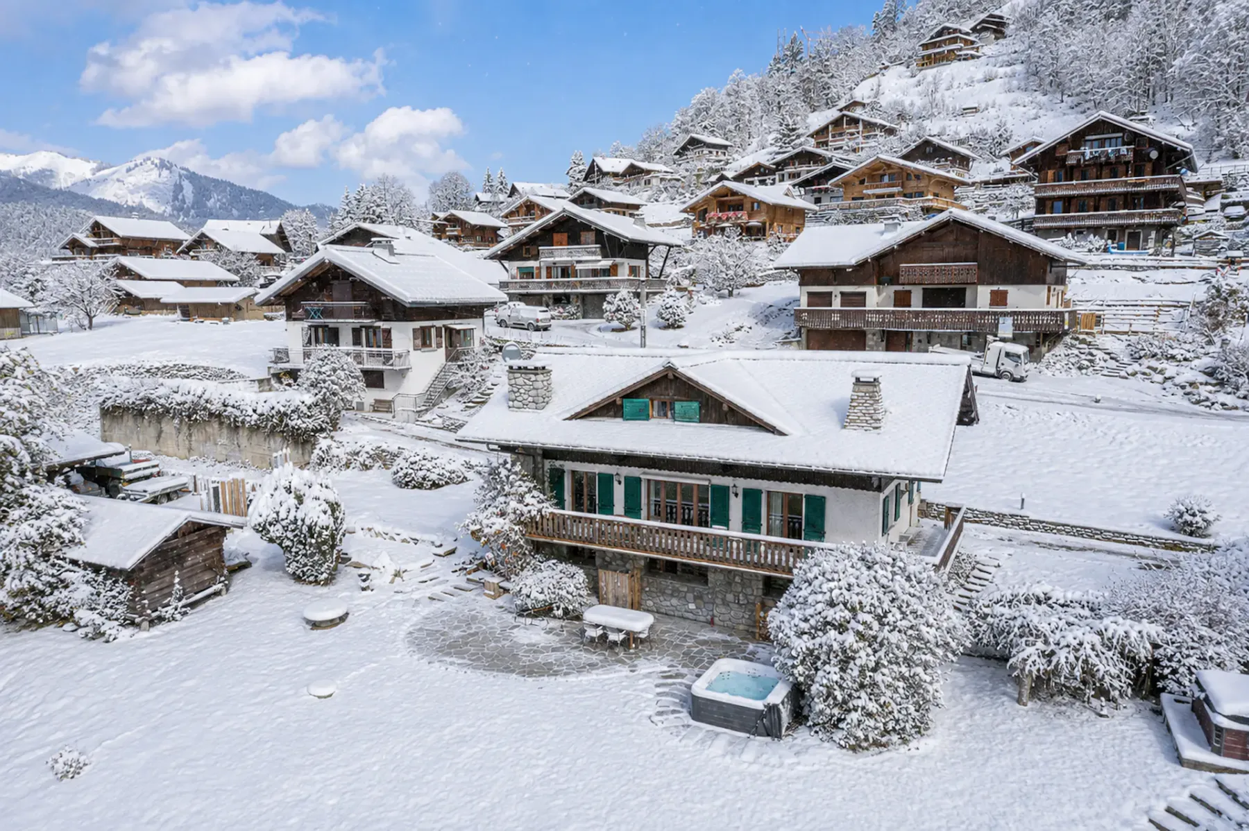 Un paisaje de pueblo nevado con chalets de madera enclavados en una ladera, rodeados de árboles cubiertos de nieve y bajo un cielo azul con nubes.
