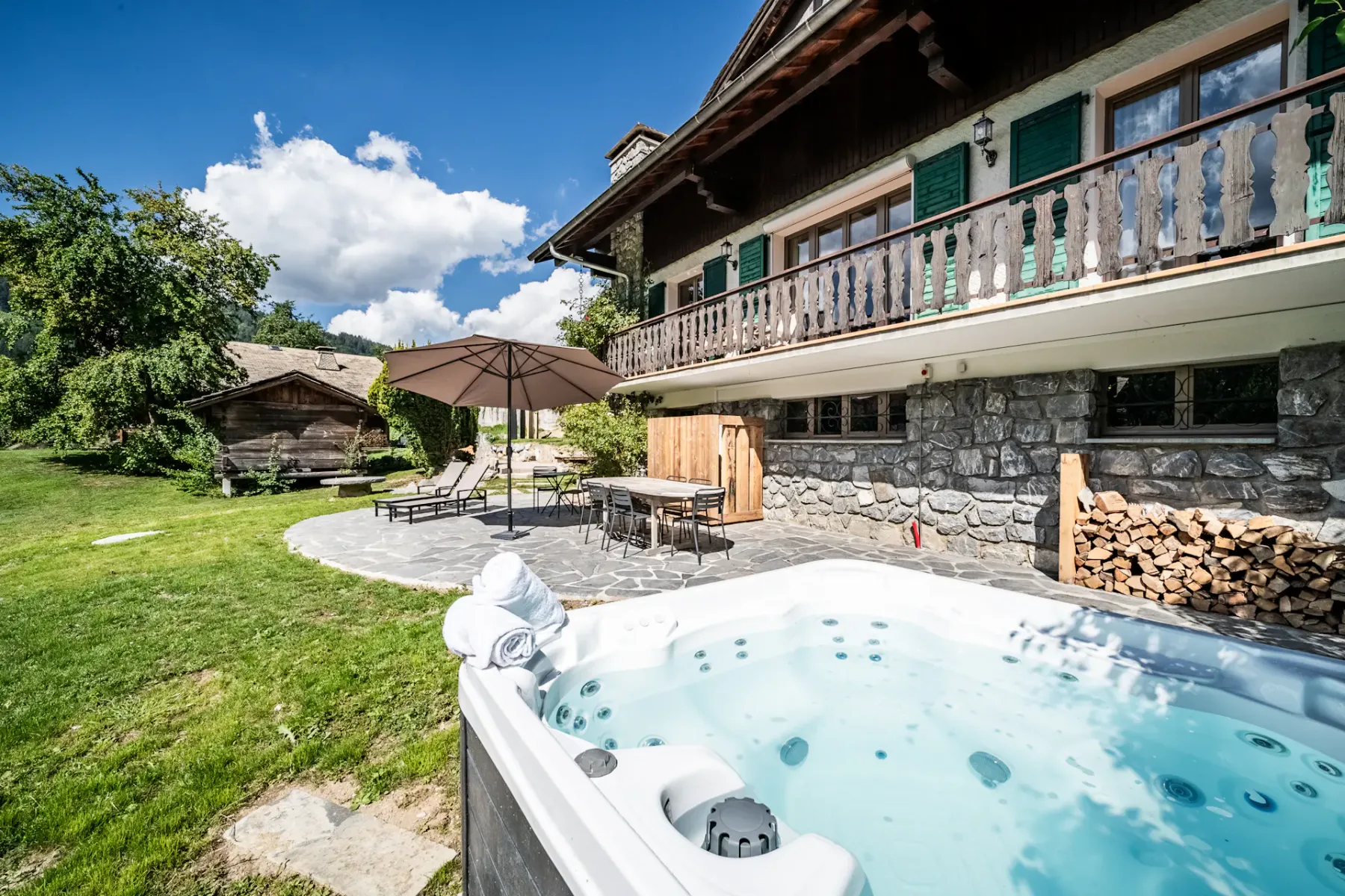 Encantador chalet de madera con bañera de hidromasaje, muebles de jardín y césped cuidado, bajo un cielo azul con nubes algodonosas