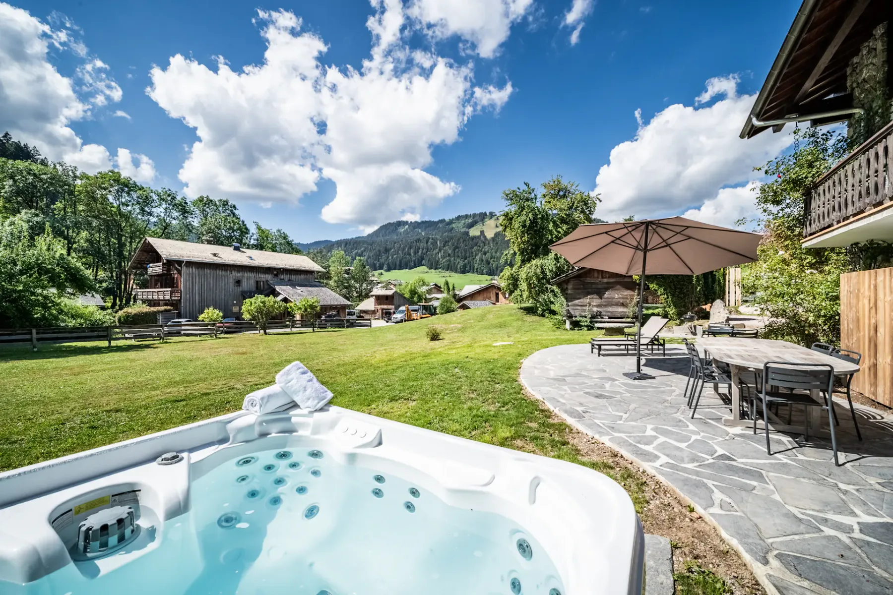 Encantador chalet de madera con bañera de hidromasaje, muebles de jardín y césped cuidado, bajo un cielo azul con nubes algodonosas