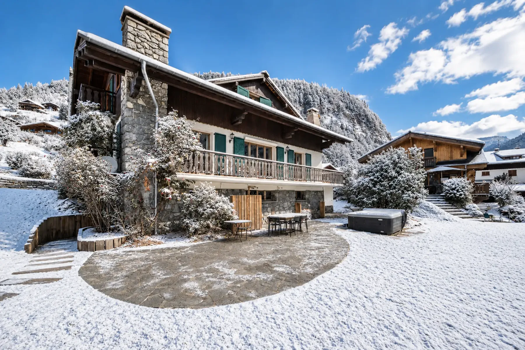 Un chalet cubierto de nieve con arquitectura de piedra y madera, rodeado de árboles y montañas nevados bajo un cielo azul despejado.