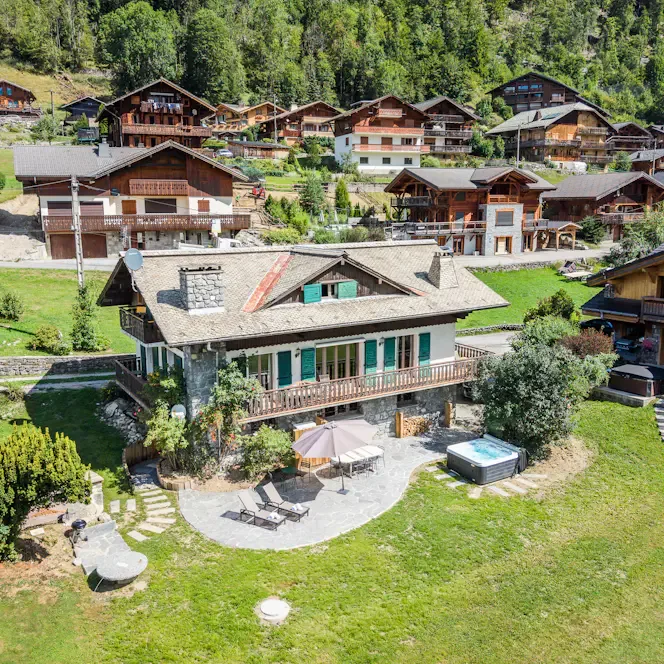 Un chalet está en un pueblo alpino verde, con un patio y asientos al aire libre delante.