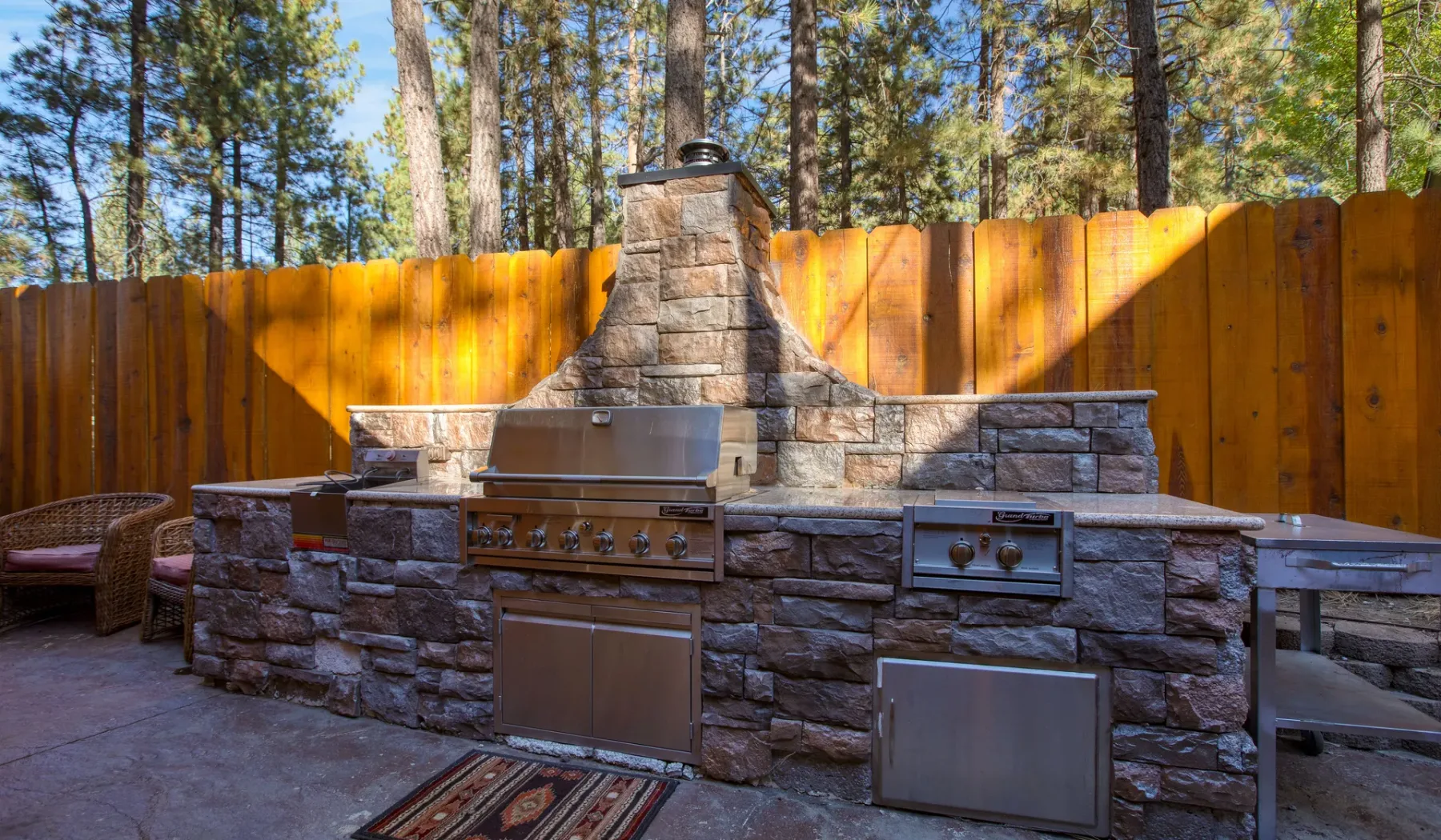 An outdoor stone cooking area stands against a wooden fence among tall trees.