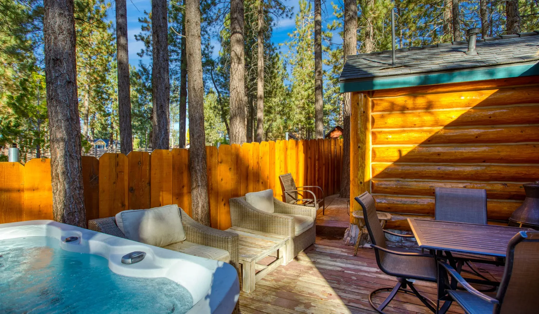 A forest-side deck features an outdoor hot tub, seating, and a wooden table.