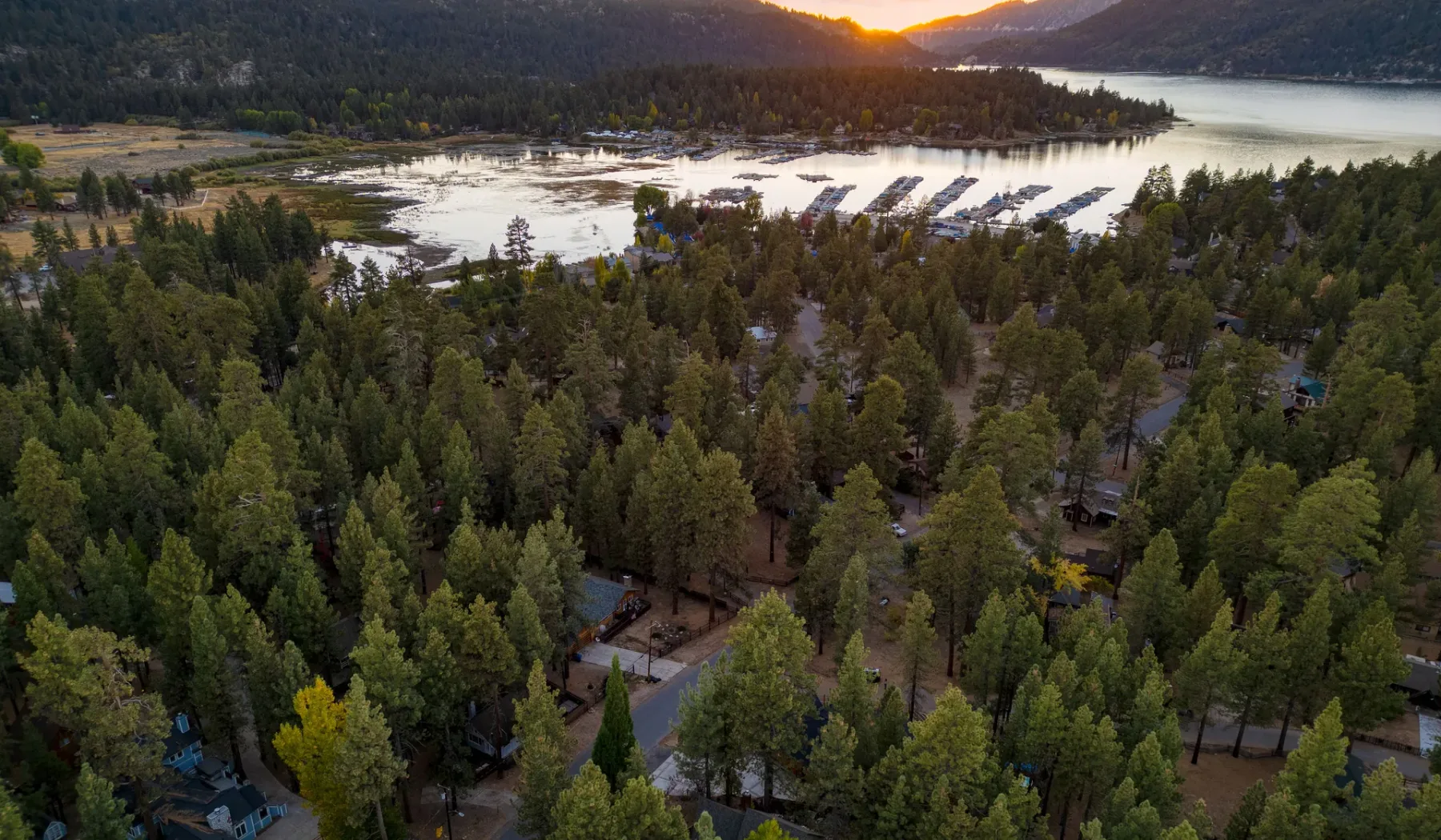 A forested lakeshore and campground sit below mountains at sunset.