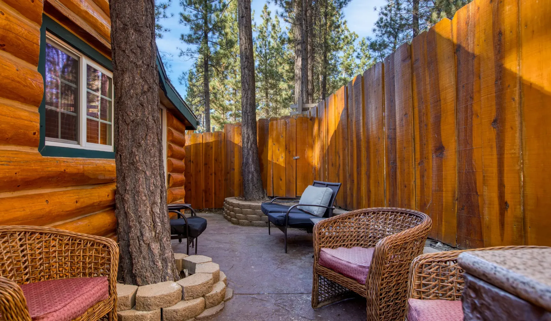 An outdoor patio with wicker seating sits beside a log cabin and wooden fence.