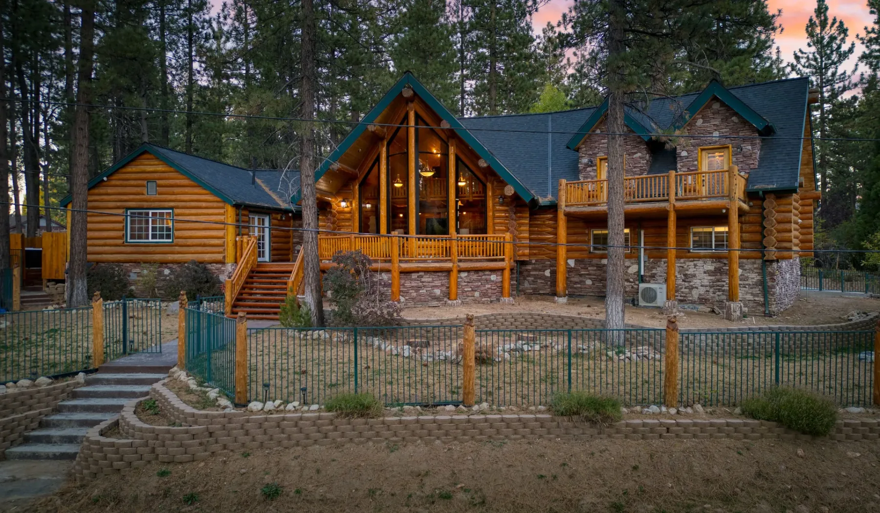 A large log cabin with a peaked entrance stands among tall trees.