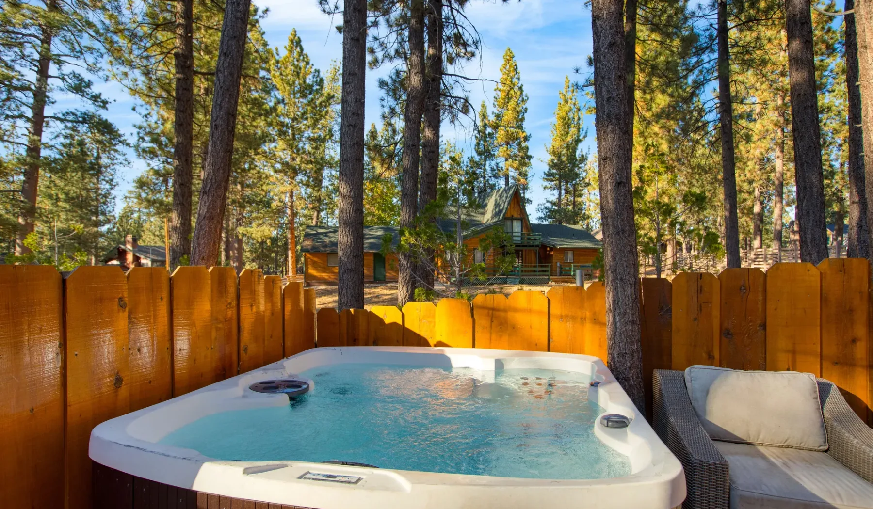 An outdoor hot tub sits beside wooden fencing among tall trees.