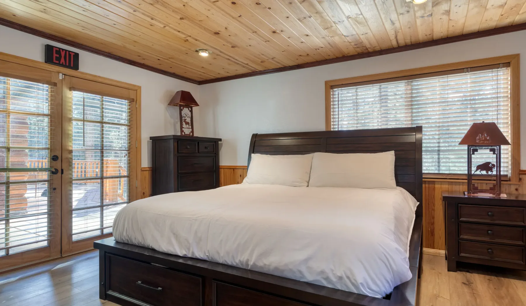 A bedroom with a large bed, wood-paneled ceiling, and windows with blinds.