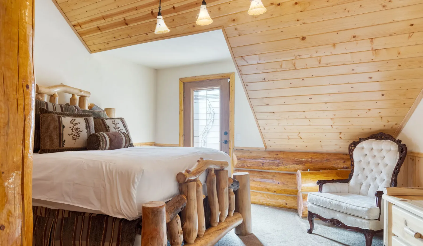 A rustic bedroom with a raised bed and an upholstered chair beneath a sloped wood ceiling.