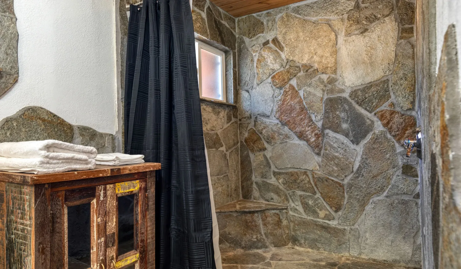 A stone-walled bathroom with a small window and folded towels on a wooden cabinet.