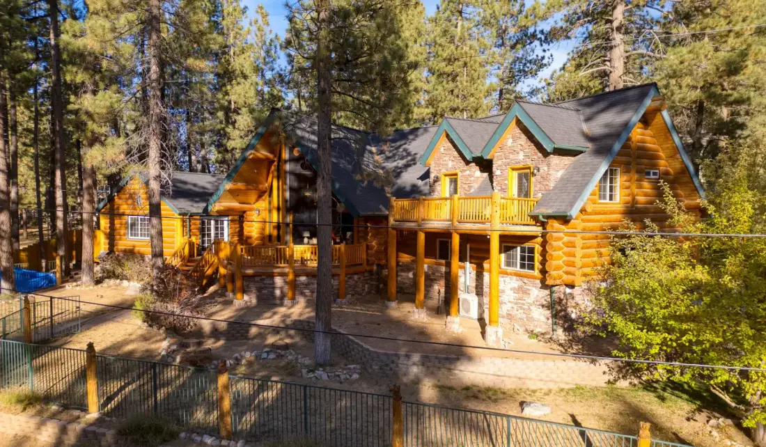 A large log cabin sits among tall pine trees, with a raised wooden deck.